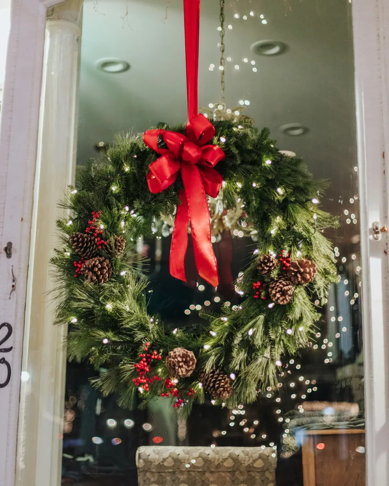 Guirlanda volumosa com pinhas, frutinhas e laço vermelho pendurada em uma porta de vidro, com luzes de Natal ao fundo